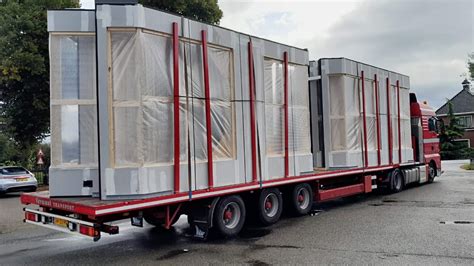 Illustratie van een grote kraan die een prefab module op een vrachtwagen laadt, met een vrachtwagen die klaarstaat voor transport.